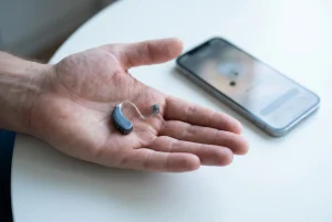 A modern slim digital hearing aid in a dark slate colour held in an open palm, beside a smartphone in soft focus on a clean white surface, cool daylight, hinting at wireless connec